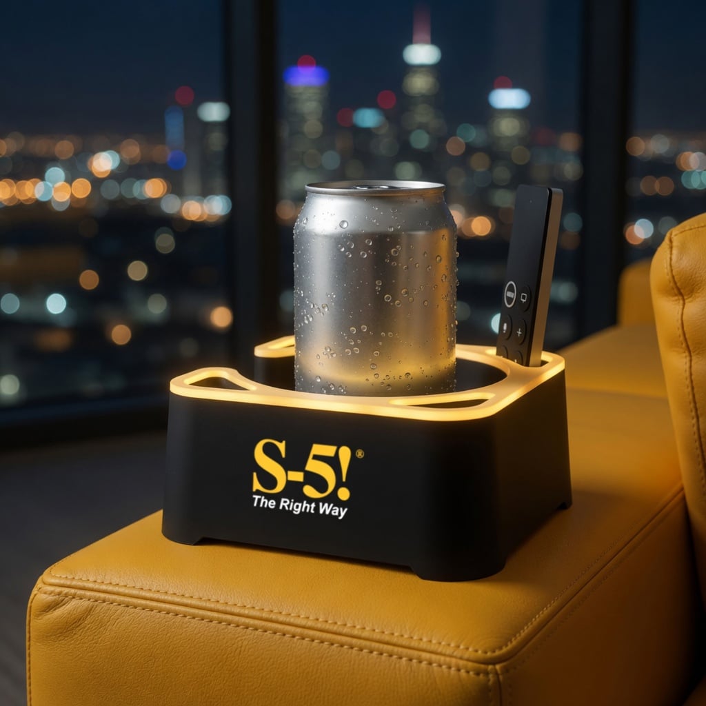 Black MugBrick Glow with a yellow glow ring and an S-5! logo holding a soda can and a TV remote on a saffron leather sofa arm, with city lights at night behind